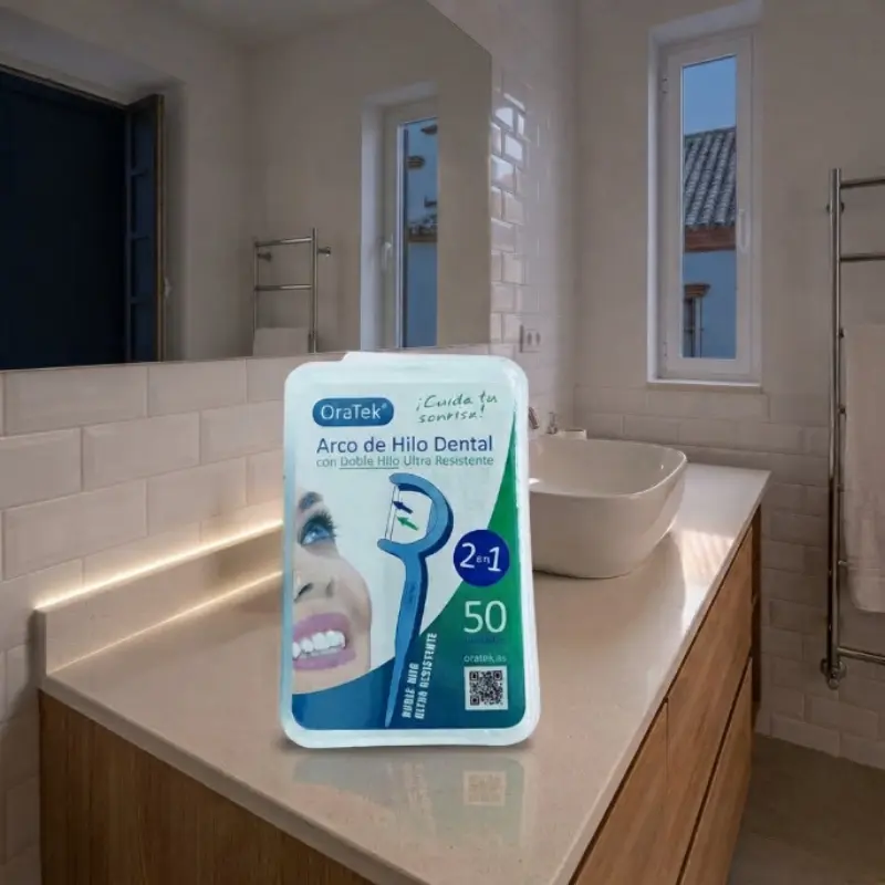 Caja de hilo dental con arco OraTek y estuche sobre el mueble de un baño iluminado con lavabo y espejo.