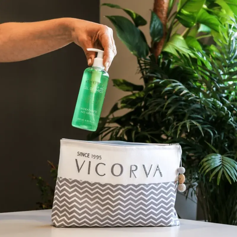 Mano introduciendo un envase de gel dérmico de aloe vera VICORVA dentro de un neceser blanco y gris con plantas al fondo.