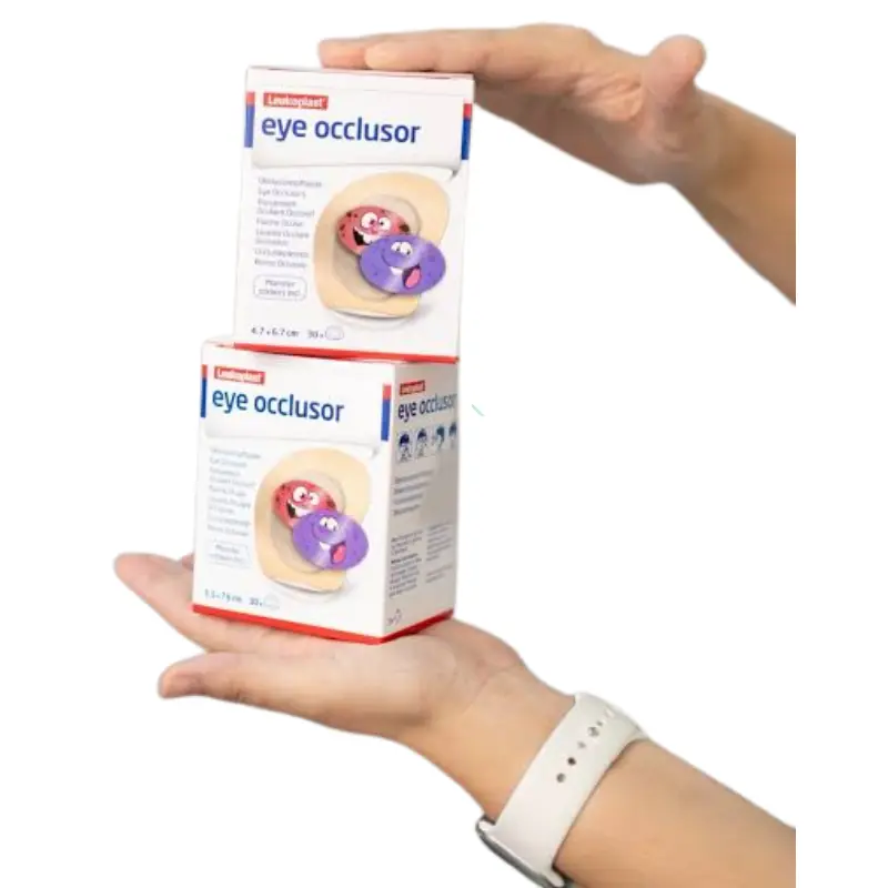 Manos sosteniendo dos cajas de parches oculares infantiles Leukoplast Eye Occlusor sobre un fondo blanco.