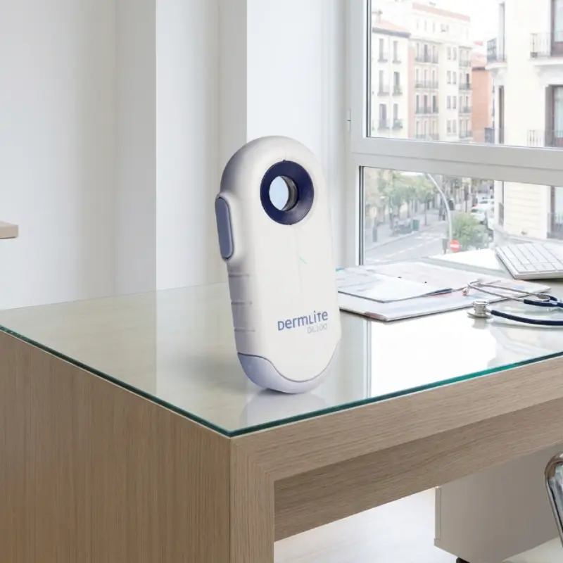 Dermatoscopio Dermlite DL100 de luz polarizada sobre una mesa de cristal en una consulta médica junto a un gran ventanal.