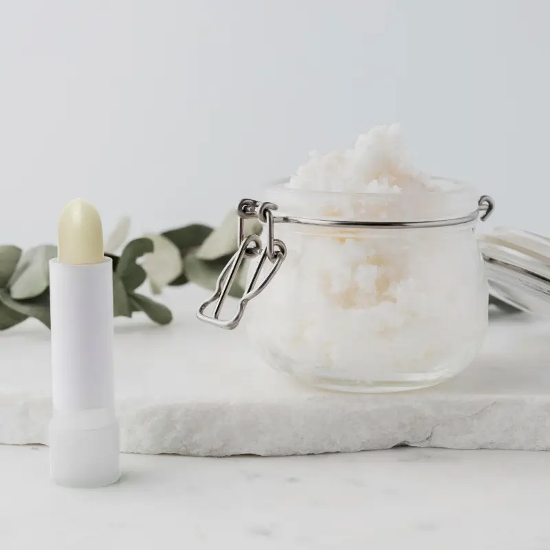 Barra de protector labial con manteca de karité colocada junto a un tarro de cristal con ingredientes naturales y hojas sobre superficie de piedra blanca.