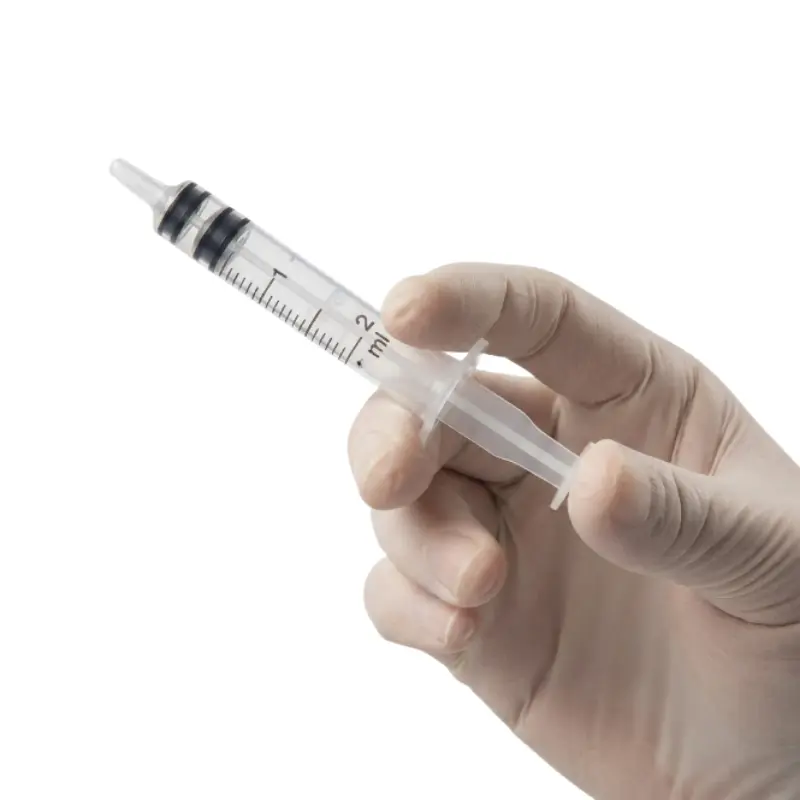 Mano con guante clínico sosteniendo una jeringa de 3 cuerpos de 2 ml Klmed con graduación visible sobre fondo blanco.