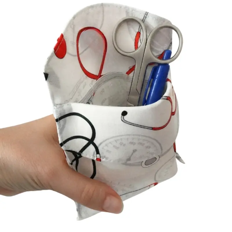 Mano sosteniendo un salvabolsillos sanitario blanco con estampado de constantes vitales, tijeras y bolígrafo azul.