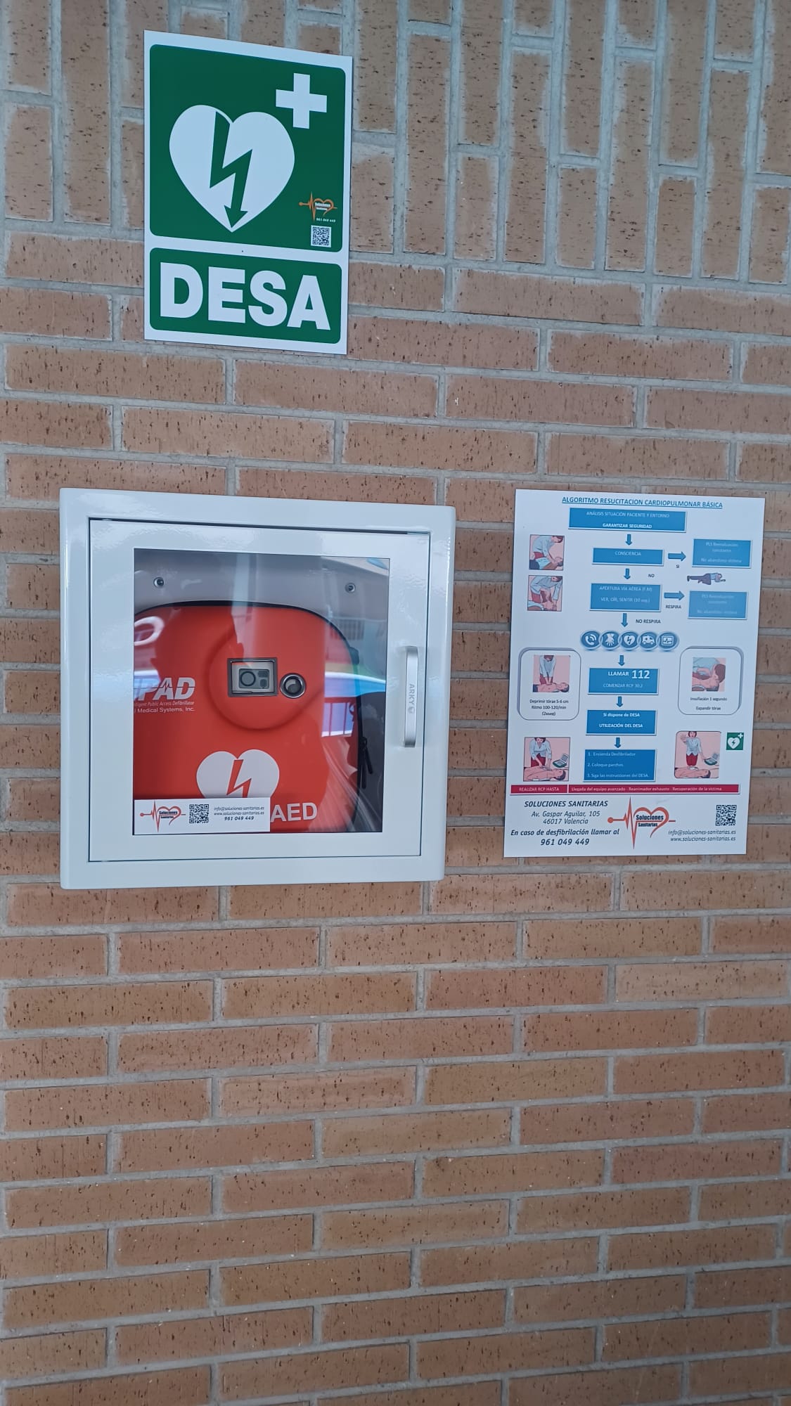 Un desfibrilador externo automático (DEA) de color rojo brillante, visible a través de una caja de pared de metal blanca con un frente de vidrio transparente. El DEA está montado sobre una pared de ladrillos, junto a una señal verde y blanca que muestra el símbolo de un corazón con un rayo y la palabra "DESA", y un póster informativo sobre reanimación cardiopulmonar (RCP) básica.
