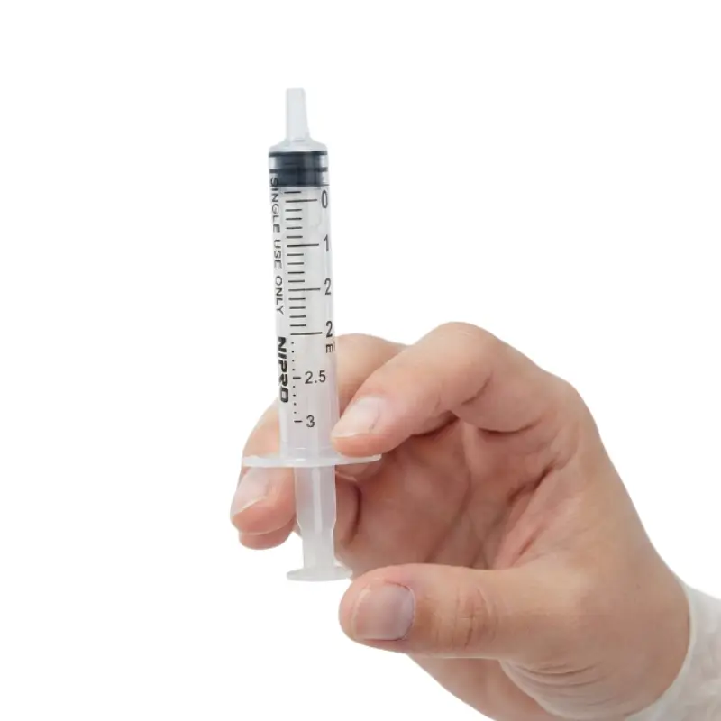 Mano enguantada sosteniendo una jeringa transparente Nipro de 2 ml con sistema Luer Lock sobre fondo blanco.
