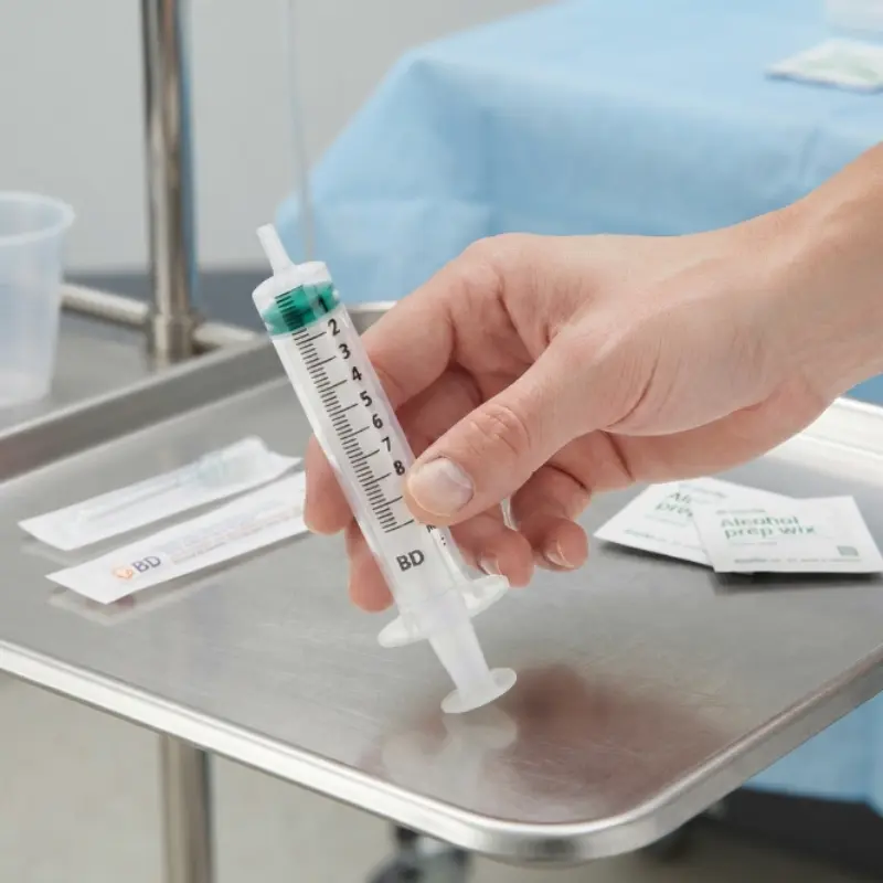 Mano sosteniendo una jeringa transparente de 3 cuerpos BD de 10 ml sobre una bandeja clínica de acero inoxidable con gasas.