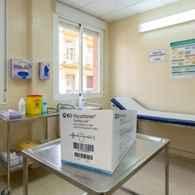 Caja de palomillas BD Vacutainer Safety-Lok situada sobre una mesa metálica en una sala de extracciones clínicas con camilla al fondo.