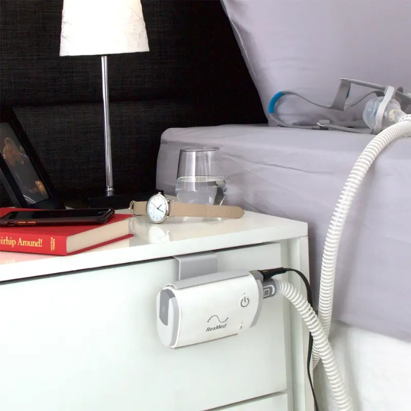 Dispositivo CPAP AirMini montado en el lateral de una mesita de noche blanca junto a una cama con ropa gris.