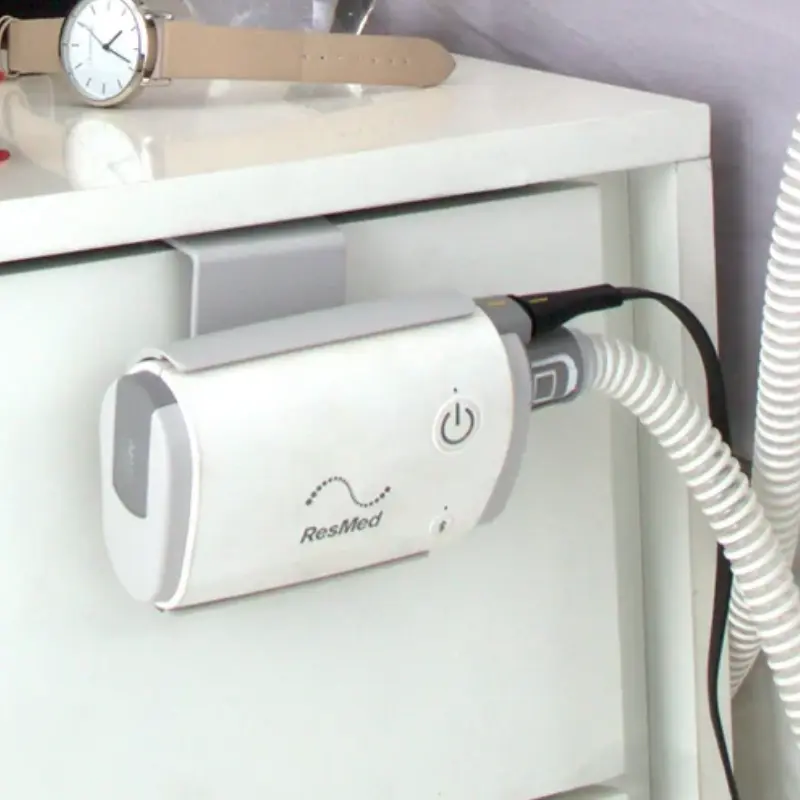 Detalle del dispositivo CPAP AirMini montado con su soporte lateral en el borde de una mesita de noche blanca.