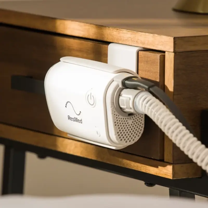 Detalle del soporte lateral del dispositivo CPAP AirMini ResMed instalado en el borde de un cajón de madera.
