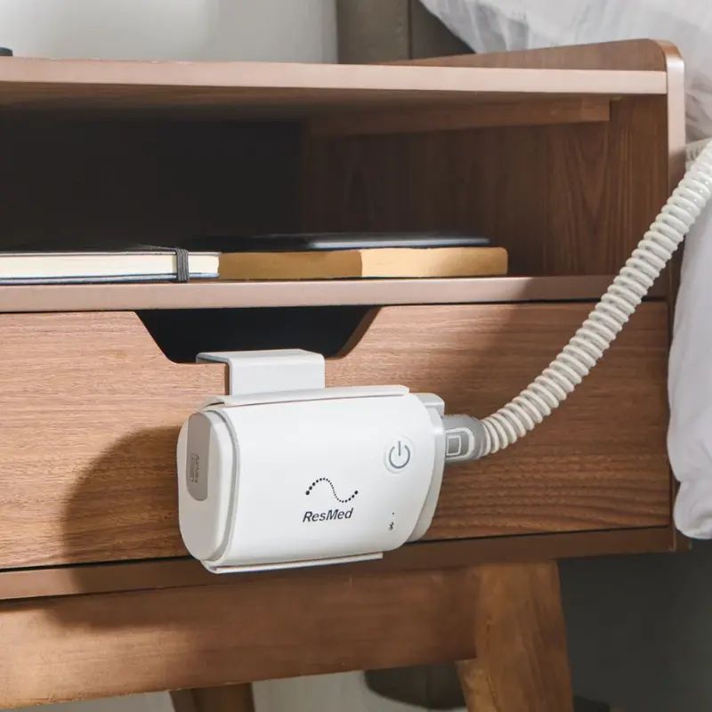 Dispositivo CPAP AirMini sujeto en el lateral de una mesita de noche de madera con libros en la balda superior.