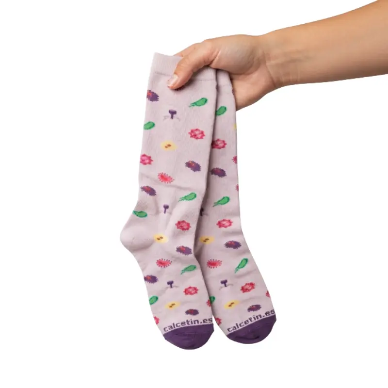 Mano sosteniendo un par de calcetines compresivos de color claro con diseño de microorganismos y bacterias.