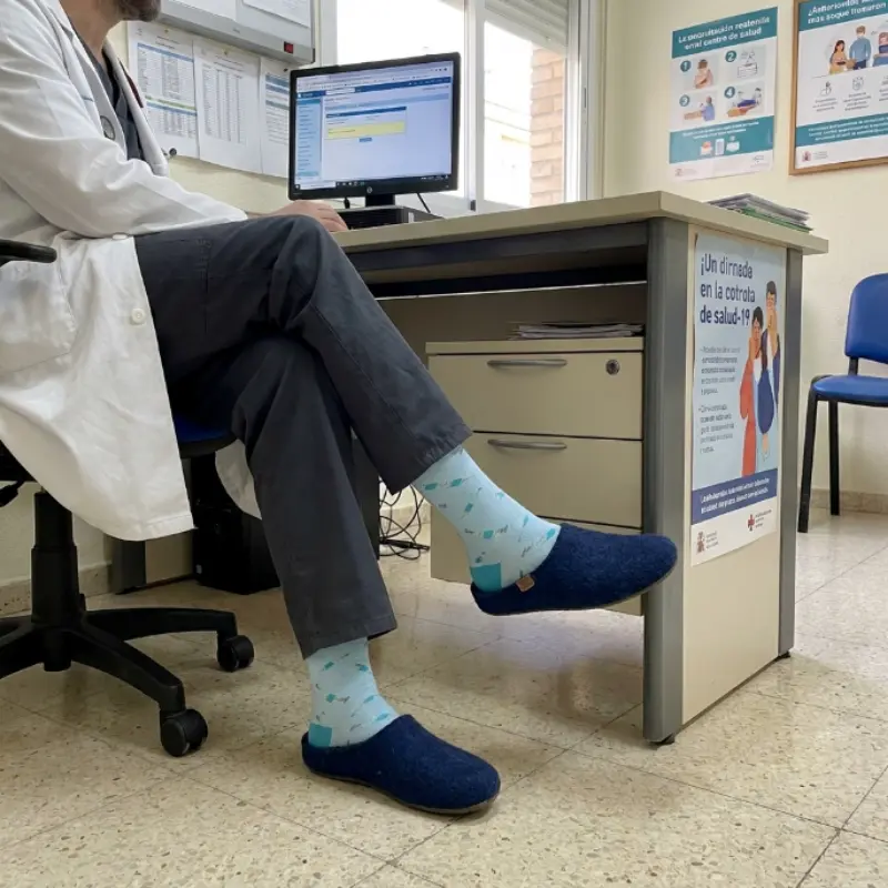 Sanitario sentado en una consulta con bata blanca y los calcetines compresivos de jeringas frente al ordenador.