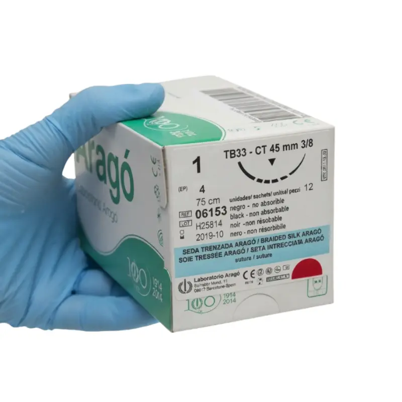 Mano con guante clínico azul sujetando una caja de sutura no absorbible de seda Aragó frente a fondo blanco.