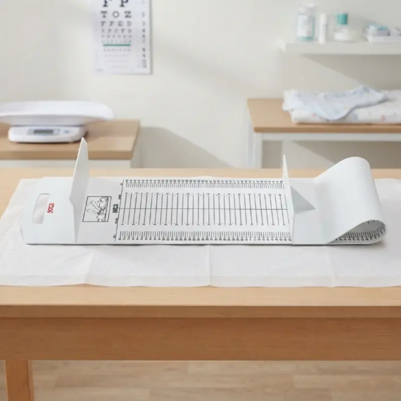 Tallímetro portátil SECA 210 extendido sobre papel de camilla en una mesa de consulta pediátrica con mobiliario al fondo.