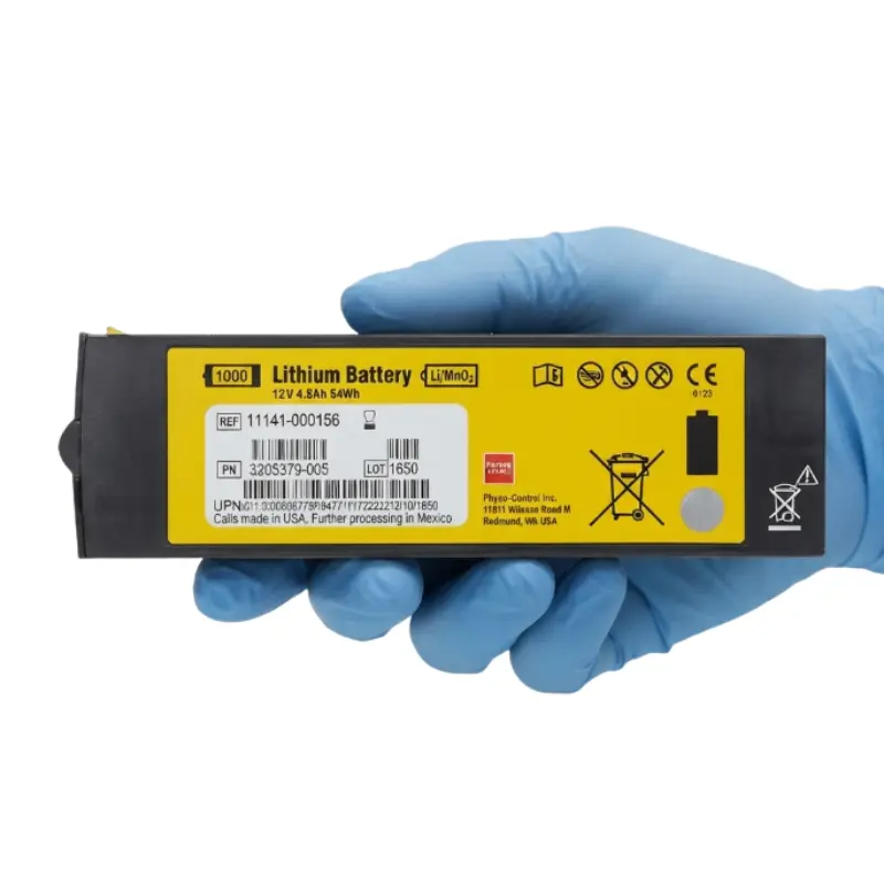 Mano con guante azul de nitrilo sosteniendo la batería de litio Physio-Control Lifepak 1000 con etiqueta amarilla informativa.