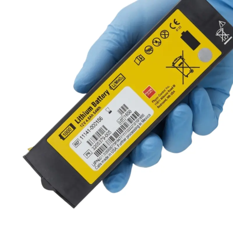 Mano con guante azul de nitrilo sosteniendo la batería de litio Physio-Control Lifepak 1000 con etiqueta amarilla informativa.