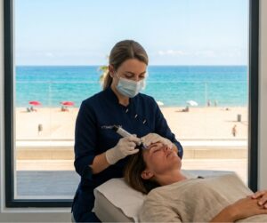 Tratamiento de mesoterapia con Dermapen en clínica estética con vistas al mar, profesional aplicando microagujas en el rostro de una paciente.