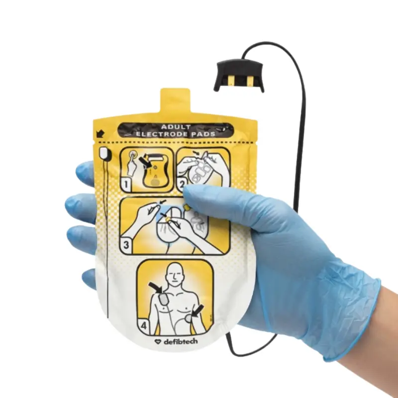 Mano con guante azul sosteniendo el envase de los electrodos de adulto Defibtech Lifeline con conector y cable.