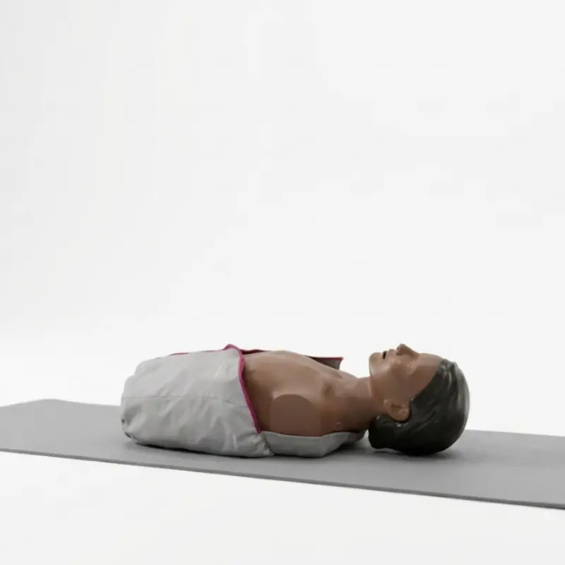 Maniquí de RCP Laerdal Little Anne QCPR de piel oscura colocado en posición horizontal sobre una esterilla gris.