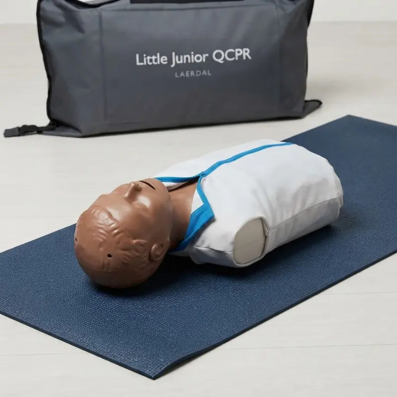 Maniquí pediátrico de RCP Laerdal Little Junior QCPR de piel oscura tendido sobre esterilla azul junto a una bolsa de transporte gris.