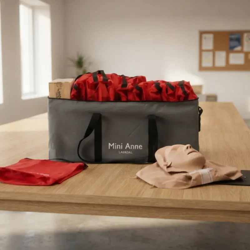 Bolsa de transporte gris abierta con múltiples estuches rojos del kit Laerdal Mini Anne Plus sobre una mesa de madera.
