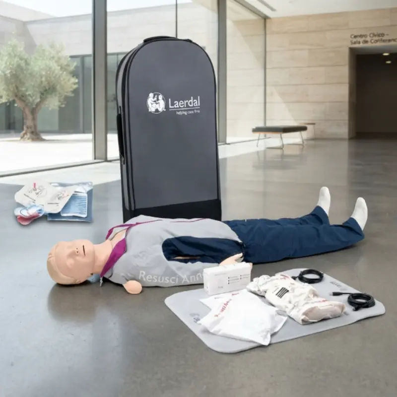 Maniquí de RCP de cuerpo entero Laerdal Resusci Anne QCPR junto a su bolsa de transporte gris y accesorios extendidos en un vestíbulo.