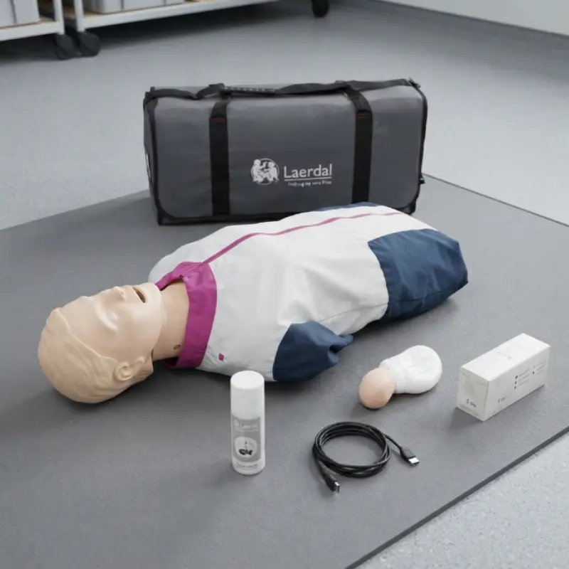 Maniquí torso de reanimación Laerdal Resusci Anne QCPR DEA junto a su bolsa de transporte y accesorios sobre alfombrilla gris.