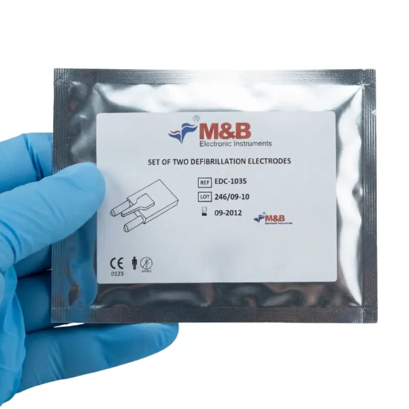 Mano con guante azul sosteniendo el envase metalizado sellado de los electrodos M&B AED 7000 sobre fondo blanco.