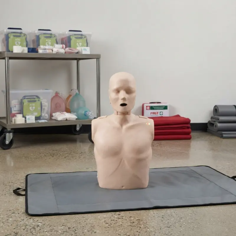 Maniquí de RCP para adultos Serie Prestan 2000 en tono claro colocado sobre una alfombrilla en una sala con estanterías y maletines médicos al fondo.