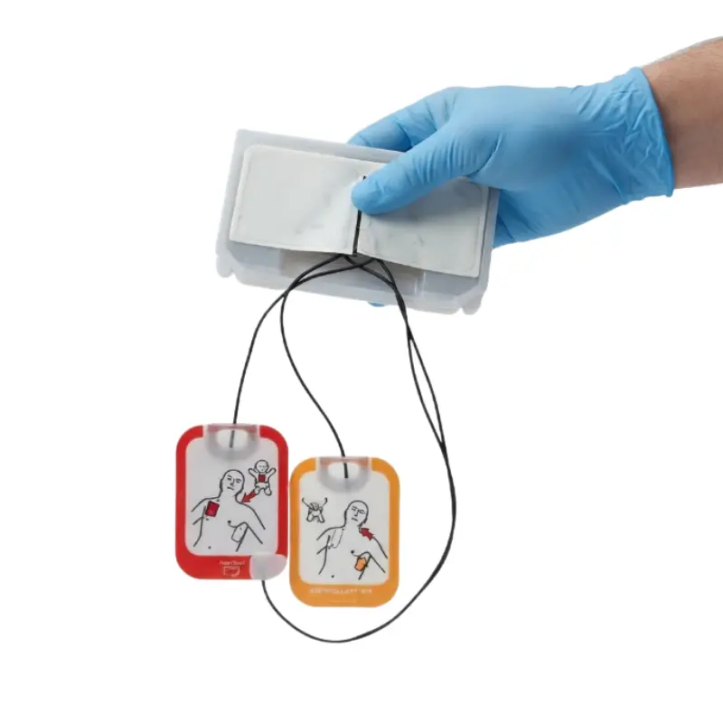 Mano con guante azul sosteniendo el cartucho de electrodos del desfibrilador Physio-Control Lifepak CR2 y sus cables.