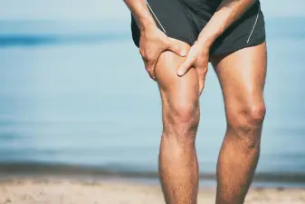 Joven sufriendo un calambre muscular en la pierna mientras pasea por la arena de la playa.