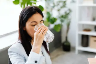 Mujer con chaqueta clara bebiendo un vaso de agua en la oficina para mantenerse hidratada y combatir las piernas cansadas.