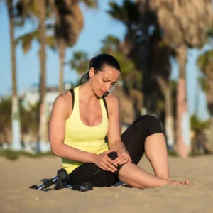 Mujer con ropa de entrenamiento amarilla sentada en la playa de arena, agarrándose la rodilla con expresión de dolor, con mancuernas cerca.