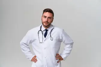 Doctor con barba, bata blanca y fonendoscopio al cuello con las manos en la cintura sobre fondo neutro.