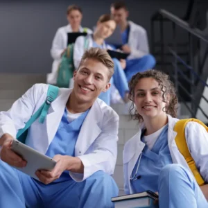 Estudiantes de medicina sonrientes con batas blancas y fonendoscopios sentados en las escaleras de la facultad con una tablet.