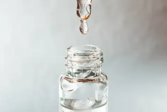 Una gota de ácido hialurónico transparente cae desde una cánula hacia la boca de un vial de vidrio estéril.