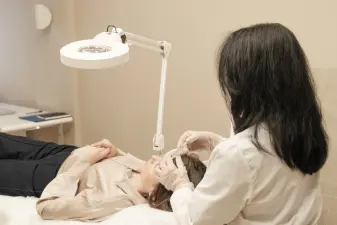 Personal sanitario con guantes aplica una inyección facial a una paciente acostada en una camilla bajo una lámpara clínica.
