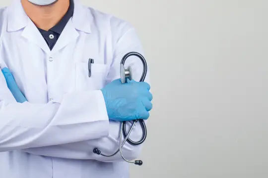 Médico con bata blanca sosteniendo un fonendoscopio en un consultorio clínico.