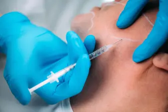 Personal con guantes azules sostiene una jeringa fina para infiltrar principios activos en el rostro de un paciente.