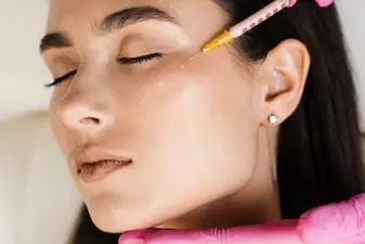 Especialista con guantes rosas aplicando una infiltración facial con microaguja en el pómulo de una paciente.