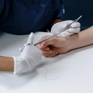 Manos con guantes blancos utilizando pinzas y un porta-agujas para realizar una sutura sobre la piel en un entorno clínico.