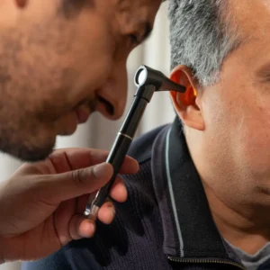 Profesional sanitario examinando el oído de un paciente adulto con un otoscopio negro equipado con luz.
