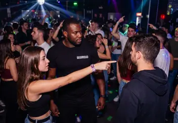 Joven señalando con el dedo a un hombre acompañada por un guardia de seguridad en una discoteca con luces de fiesta.