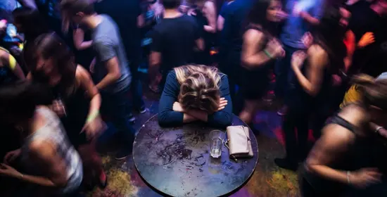 Joven con la cabeza apoyada sobre los brazos en una mesa en una discoteca con multitud de personas alrededor.