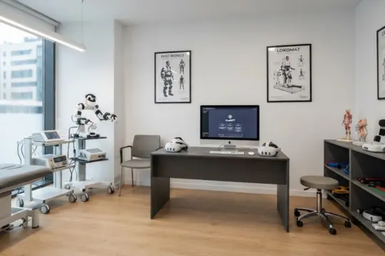 Gabinete de fisioterapia equipado con camilla, carro de electroterapia, escritorio con ordenador y carteles de biomecánica.
