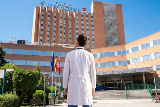 Estudiante de medicina con bata blanca y fonendoscopio de espaldas frente a la fachada del Hospital 12 de Octubre de Madrid.