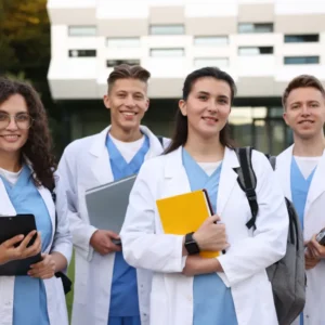 Grupo de cuatro estudiantes de medicina y enfermería sonrientes con batas blancas y pijamas azules frente a un edificio clínico moderno.