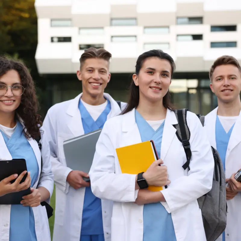 Grupo de cuatro estudiantes de medicina y enfermería sonrientes con batas blancas y pijamas azules frente a un edificio clínico moderno.