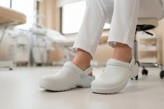 Zuecos sanitarios blancos con tira de sujeción en el talón calzados por una persona sentada en una clínica.